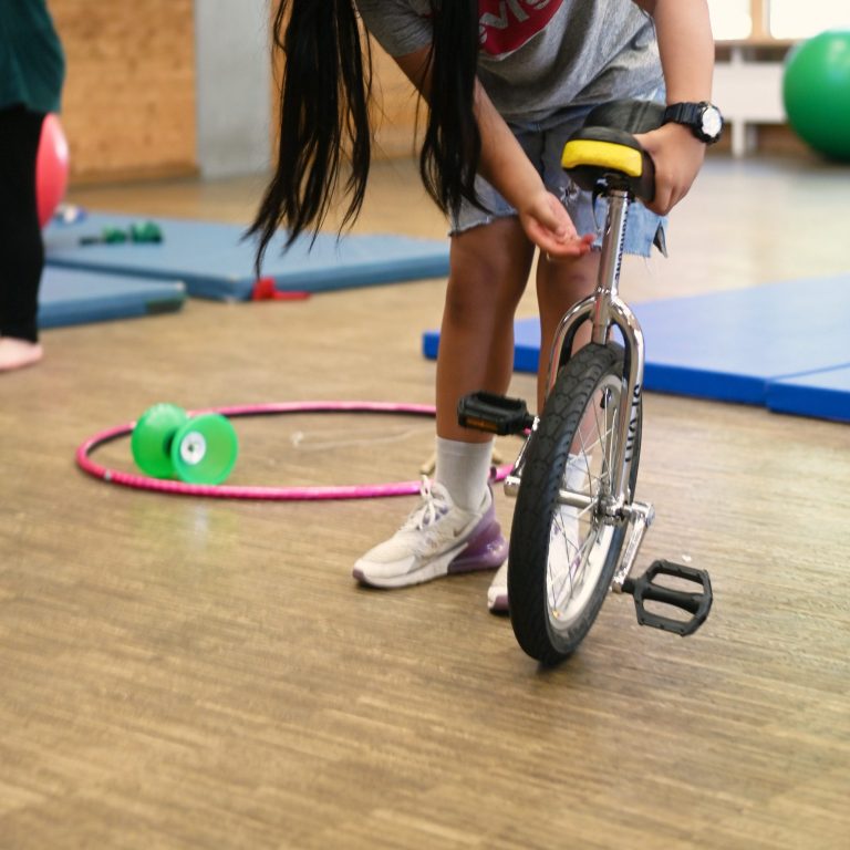 Ein Kind stellt ein Einrad auf einem Sportboden ein, im Hintergrund sind Trainingsgeräte.