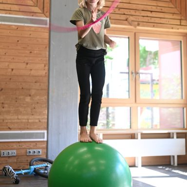 Mädchen balanciert barfuß auf einem grünen Gymnastikball und schwingt ein Seil.