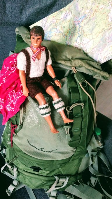 Kleine Figur im traditionellen Outfit sitzt auf einem grünen Rucksack mit einer Karte.