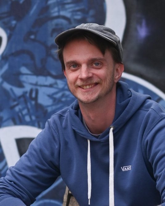 Junger Mann in blauer Mütze vor graffiti-bedeckter Wand mit dem Schriftzug „QUIBB“.