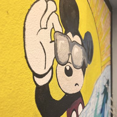 Mickey Maus mit Sonnenbrille, steht vor einer gelben Wand mit einer Welle.