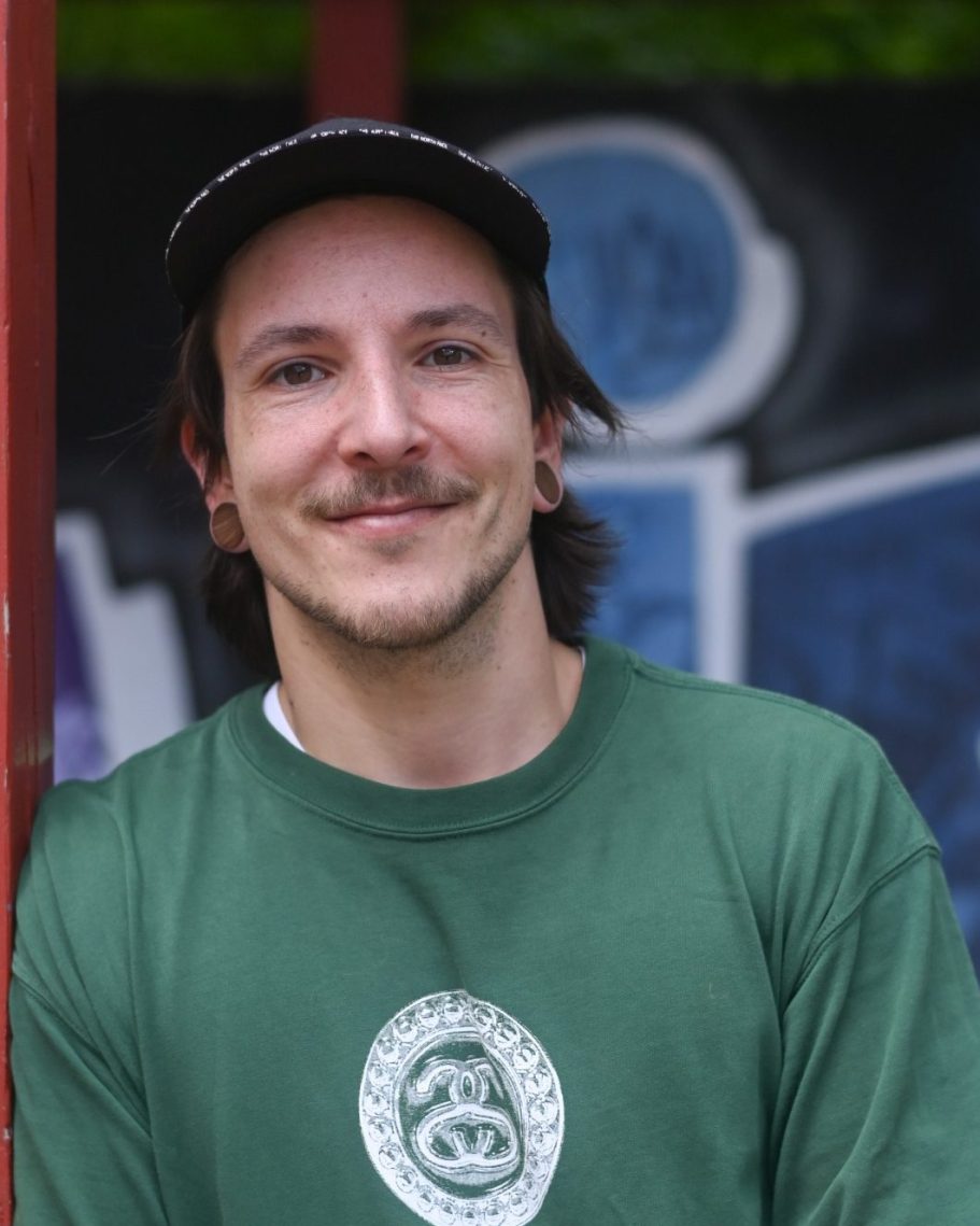 Ein junger Mann mit einem lila T-Shirt steht vor einem Wandbild mit Fischen.
