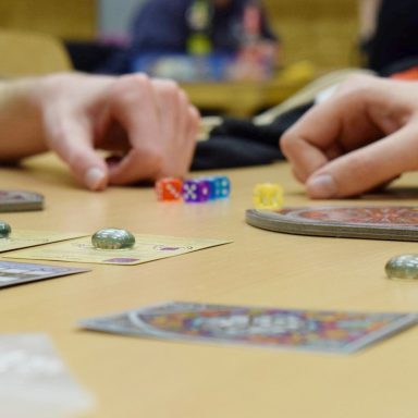 Zwei Hände spielen ein Brettspiel mit Würfeln und Spielkarten auf einem Tisch.