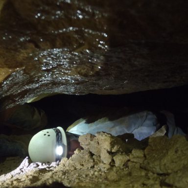 Person mit Helm, der in einer engen Höhle arbeitet, umgeben von Erde und Steinen.