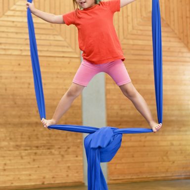 Ein Mädchen balanciert auf blauen Tüchern in einem Sportraum.