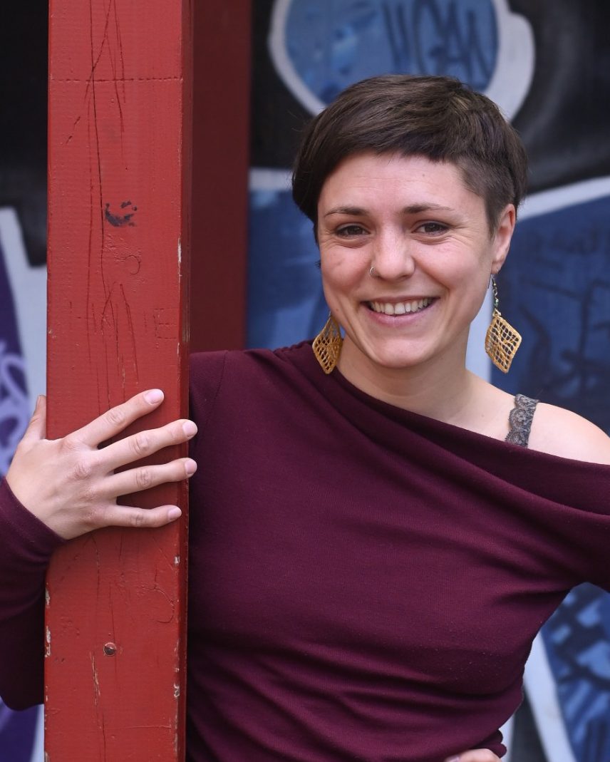 Junge Frau mit kurzen Haaren und großen Ohrringen steht an einer roten Säule.