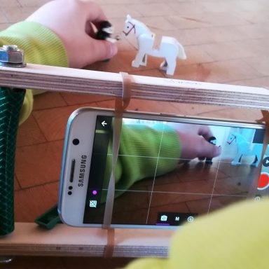 Eine Hand positioniert eine kleine Pferdefigur vor einem Smartphone auf einem Tisch.