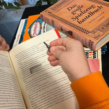 Eine Hand hält einen Lesezeichen in einem Buch, umgeben von weiteren Bücher.