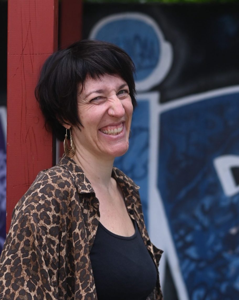 Frau mit Kurzhaarschnitt und Leopardenmuster-Bluse lächelt vor einer Graffiti-Wand.