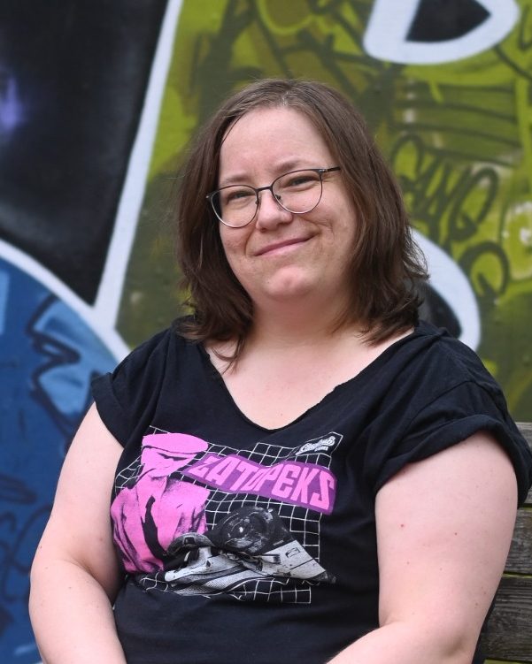 Frau mit braunen Haaren, lächelt, trägt ein schwarzes T-Shirt mit buntem Aufdruck.